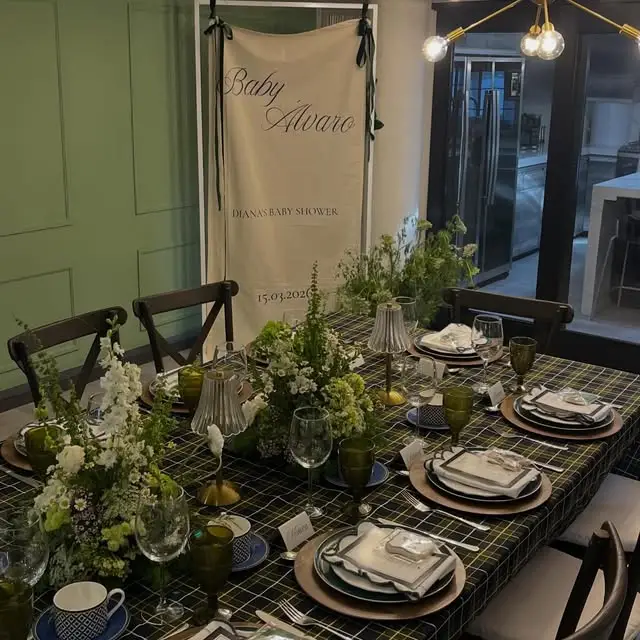 Mesa de baby shower con mantel de cuadros verde y negro, vajilla verde olivo, cristalería y arreglos de flores blancas