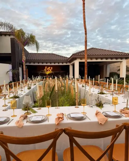 Mesa imperial larga al atardecer con mantel blanco, velas altas, servilletas color durazno y plumas de pampa entre palmeras
