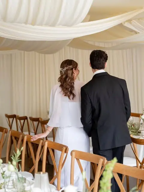 Novios de espaldas en una carpa con drapeado de tela beige en el techo y sillas cross-back de madera