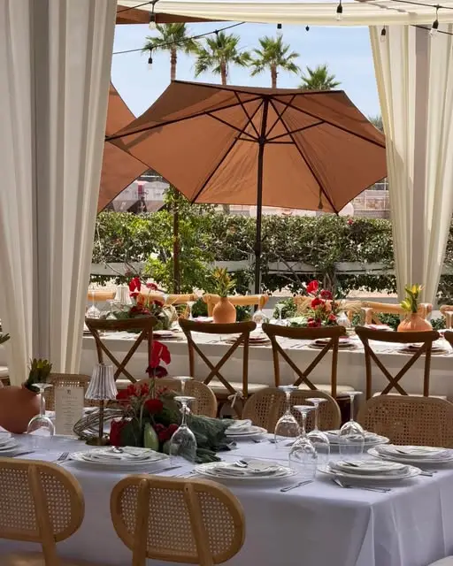 Terraza con sombrilla terracota, mesas vestidas de blanco, sillas de ratán y centros de mesa con anturios rojos