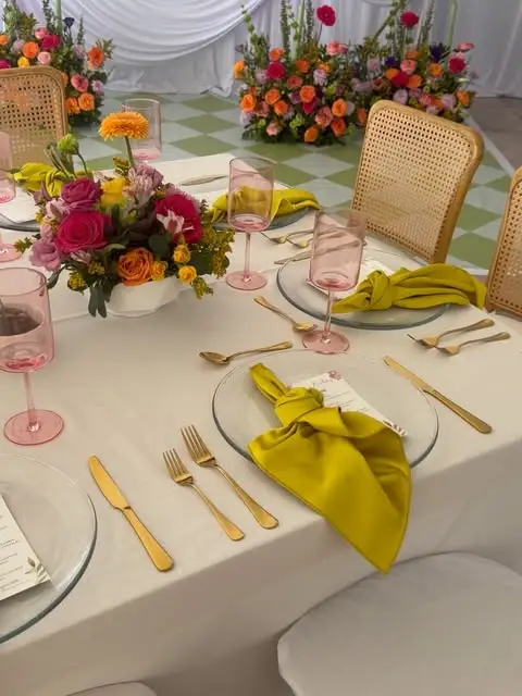 Mesa con servilletas amarillo mostaza, copas rosas, cubiertos dorados y centro floral de rosas fucsia y naranjas frente a un muro de flores